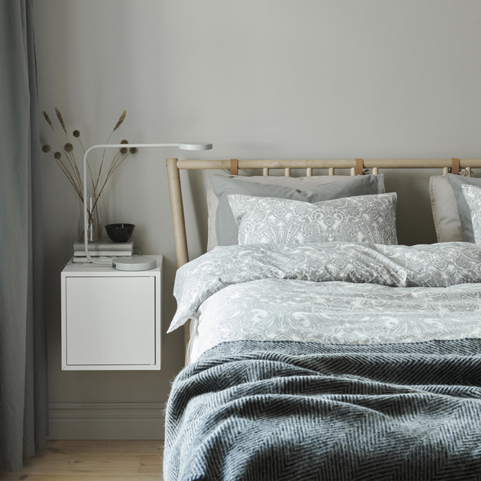 Спалня с легло с чаршафи в неутрални цветове и монтирано на стената бяло нощно шкафче.