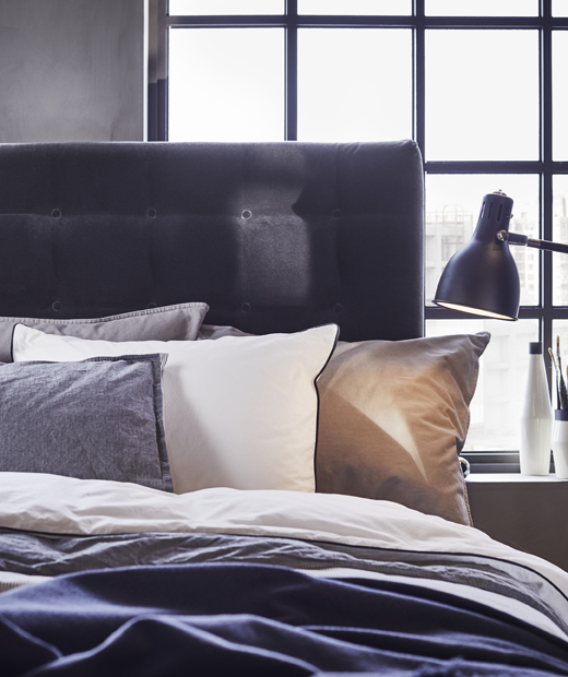 Използвай спалното бельо IKEA BLÅVINDA от 100% памук с възглавници и завивки за лспалня с усещане на хотелски лукс. Приближен изглед на легло с натрупани възглавници и завивки в бяло и сиво, до черна нощна лампа.