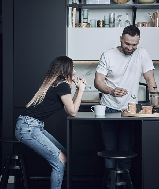 Черна кухня IKEA KUNGSBACKA с приветлив кухненски остров. Катарзина седи на бар стол до черен кухненски остров, където Миколай приготвя кафе.