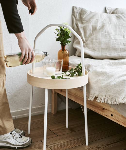 Преносимата масичка IKEA BURVIK може да се използва и като нощна масичка. Ивет сипва питие в чаша на преносима маса с бяла дръжка до легло.